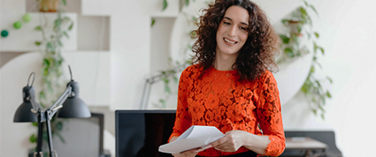 A small business owner with long hair and a bright orange shirt smiles while reviewing paperwork in their office.