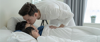 A person kisses his husband goodbye before leaving for work while their child sleeps.