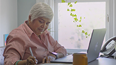 A middle-aged woman sits at a desk with an open laptop and takes notes on a pad of paper.