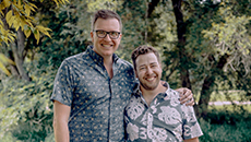 A same sex couple stands with an arm around one another in a park.