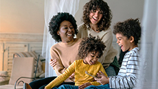 Two moms and their children laugh while one child holds a tablet computer.