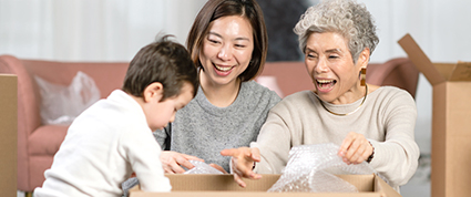 A young boy opens a moving box with his mother and grandmother who are seated on their living room floor.