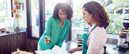 A Black TD colleague in a green sweater visits a female entrepreneur at her coffee shop to sign paperwork.