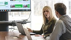 Two TD colleagues have a discussion while working on laptops with a big monitor in the background.