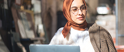 A woman wearing a hijab works on a laptop while seated outside on a Fall day.