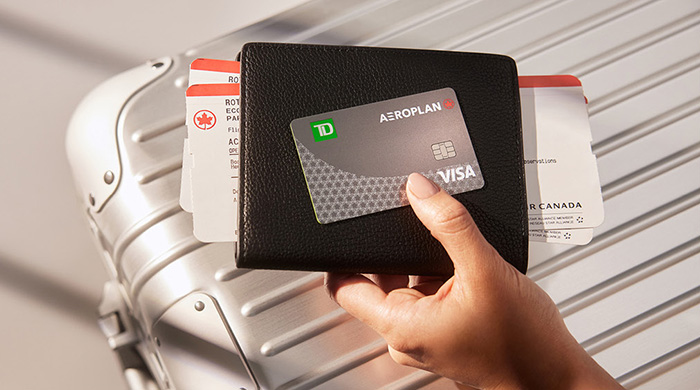 Hand holding passport carrier, airline boarding passess and a TD Aeroplan Visa card over a suitcase