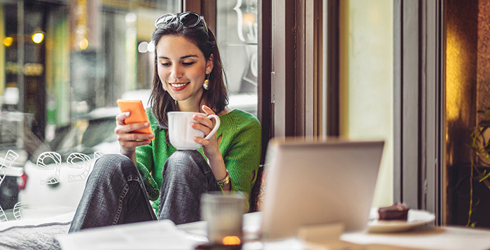 Une femme est dans un café, et tient un téléphone d’une main et une tasse de café de l’autre