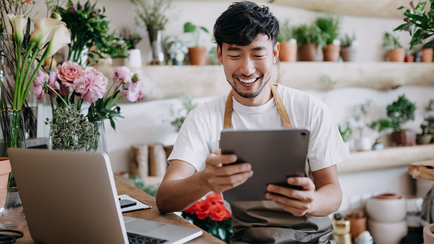 Asian male florist, owner of small business flower shop, using digital tablet while working on laptop against flowers and plants. Checking stocks, taking customer orders, selling products online. Daily routine of running a small business with technology